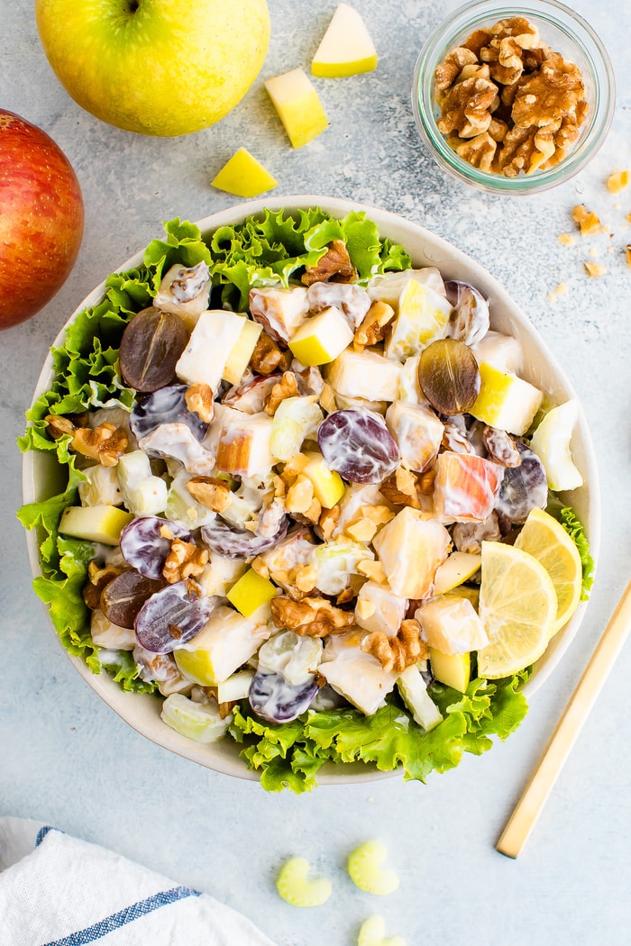 Waldorf salad served in a bowl over lettuce. Apples, walnuts and celery are around the bowl.