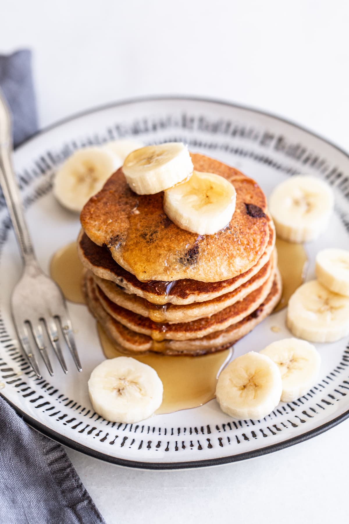 Stack of banana chocolate chip pancakes topped with banana slices and maple syrup.