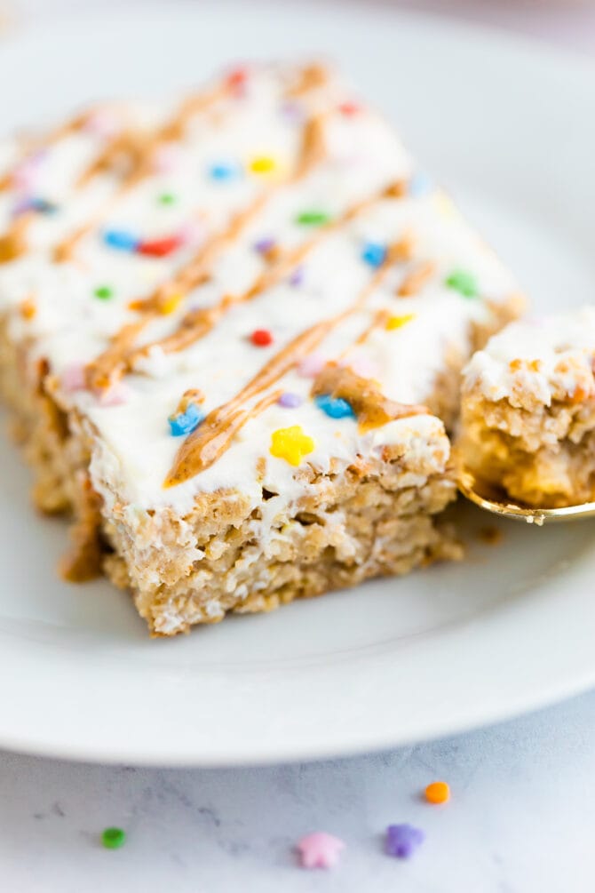 Slice of birthday cake baked oatmeal topped with frosting, nut butter drizzle and sprinkles. A spoon has taken a bite out of it.