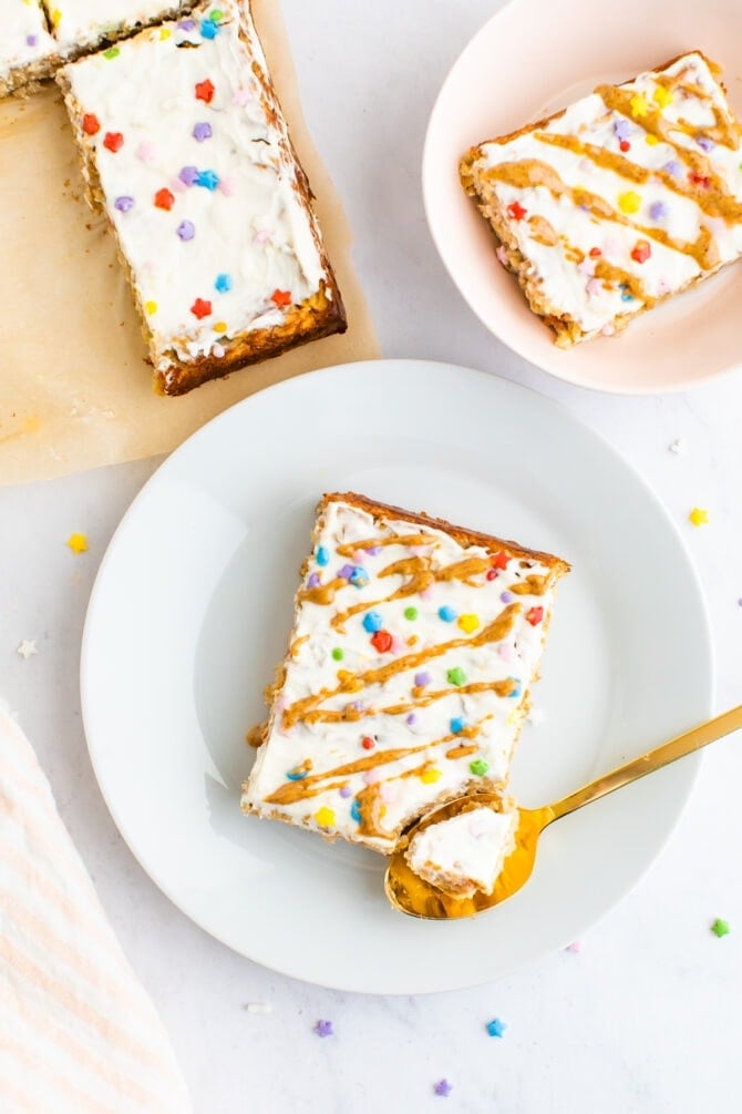 Slice of birthday cake baked oatmeal topped with frosting, nut butter drizzle and sprinkles. A spoon has taken a bite out of it.