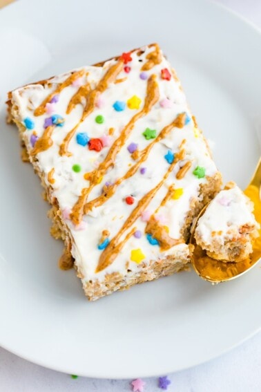 Slice of birthday cake baked oatmeal topped with frosting, nut butter drizzle and sprinkles. A spoon has taken a bite out of it.