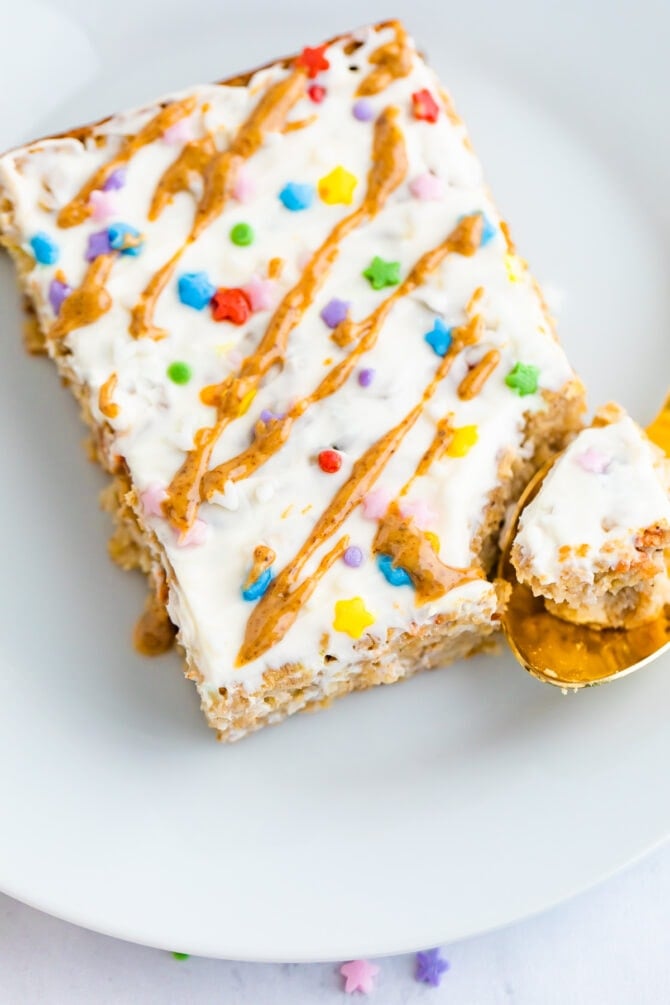 Slice of birthday cake baked oatmeal topped with frosting, nut butter drizzle and sprinkles. A spoon has taken a bite out of it.