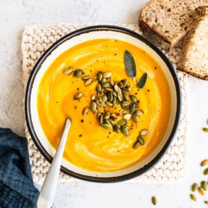 Bowl of butternut squash topped with sage, pepper and pumpkin seeds. Bread is beside the bowl.