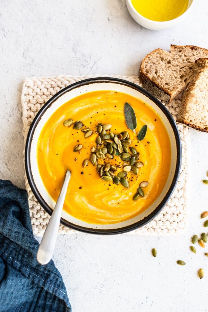 Bowl of butternut squash topped with sage, pepper and pumpkin seeds. Bread is beside the bowl.