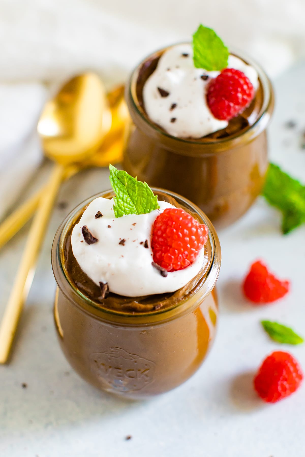 Two jars of avocado chocolate pudding topped with whipped cream, raspberries chocolate shavings and mint.