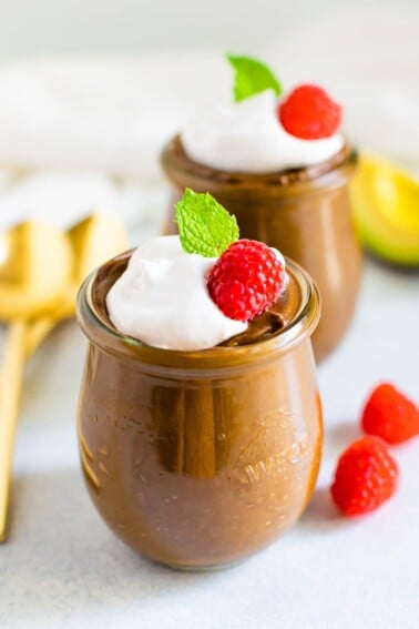 Two jars of avocado chocolate pudding topped with whipped cream, raspberries and mint.