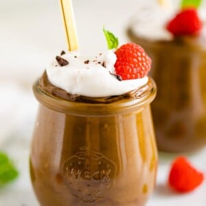 Two jars of avocado chocolate pudding topped with whipped cream, raspberries chocolate shavings and mint.