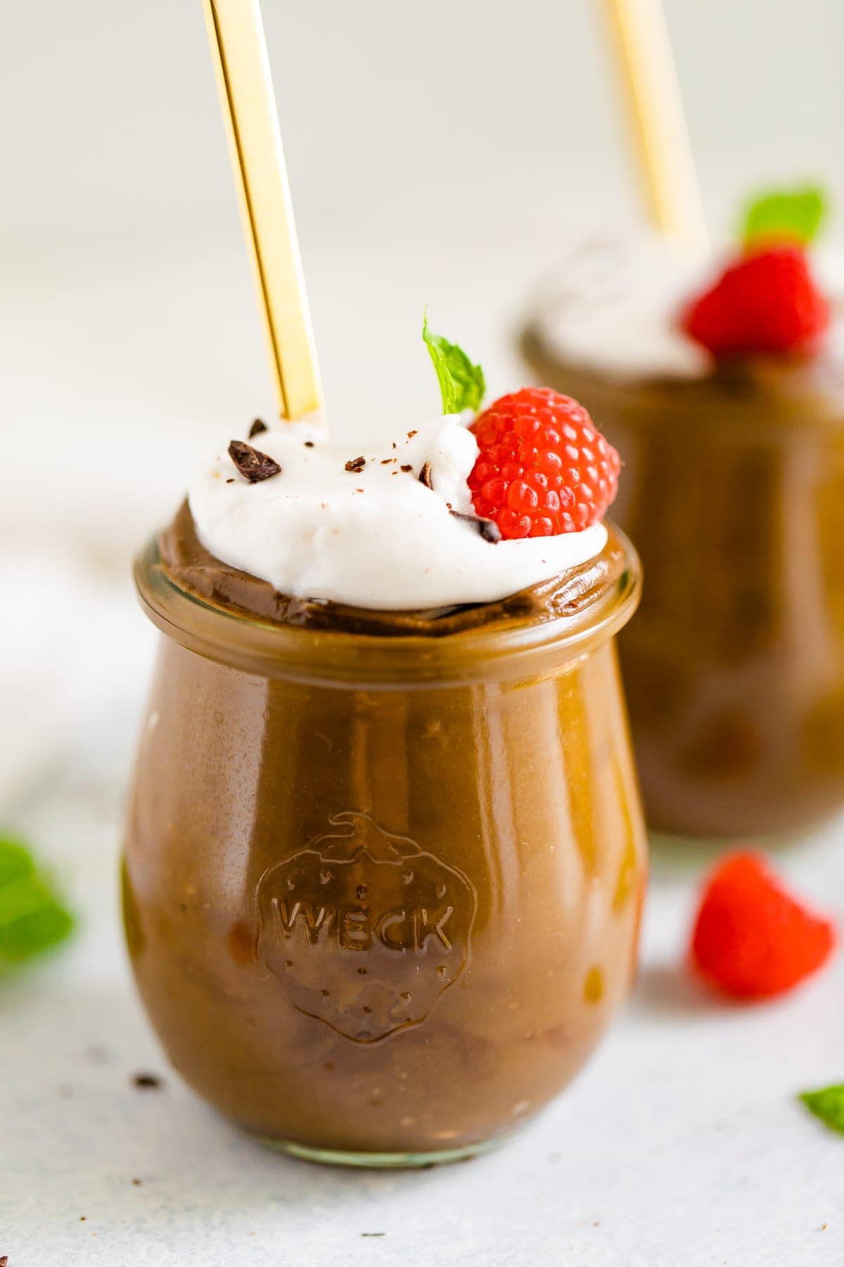 Two jars of avocado chocolate pudding topped with whipped cream, raspberries chocolate shavings and mint.