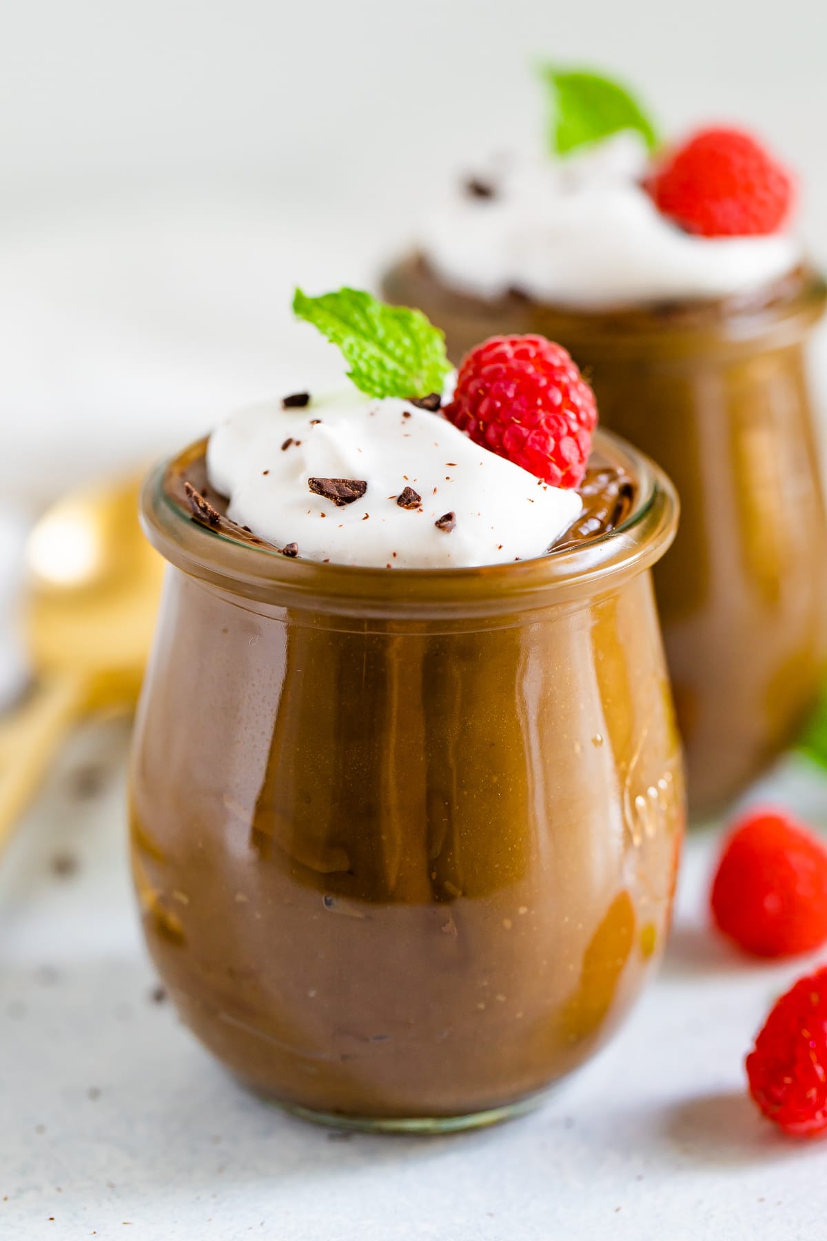 Two jars of avocado chocolate pudding topped with whipped cream, raspberries chocolate shavings and mint.