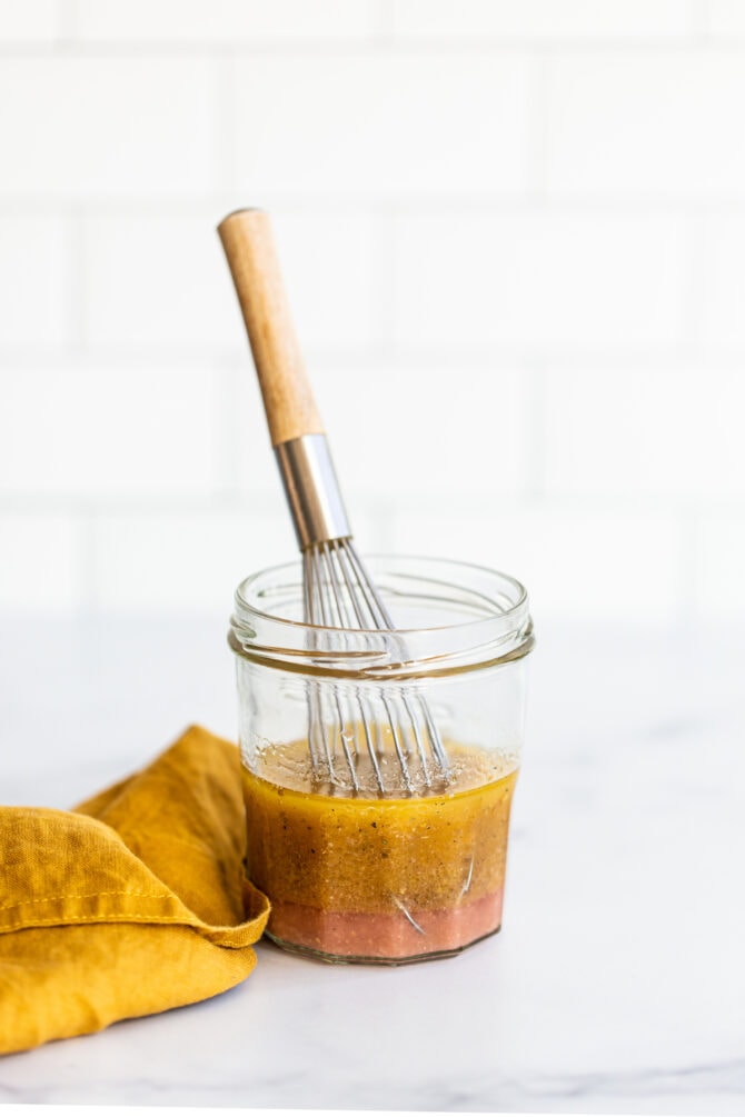 Jar of greek salad dressing with a little whisk. Yellow napkin is beside the jar.