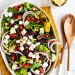 Oval platter with a greek salad. Salad tongs and lemon wedges are beside the platter.