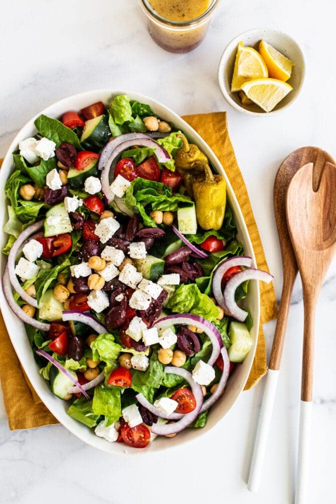 Oval platter with a greek salad. Salad tongs and lemon wedges are beside the platter.