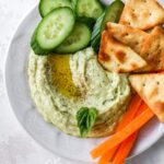 Bowl of lima bean hummus garnished with olive oil, pepper and basil and served with cucumbers, carrots and crackers.