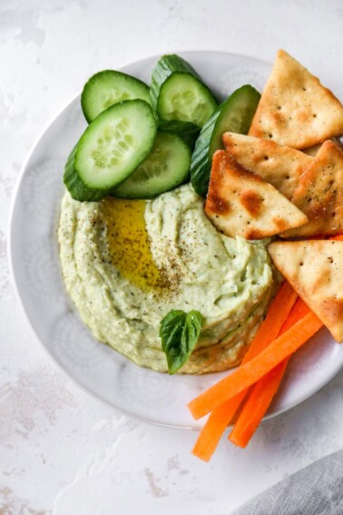 Bowl of lima bean hummus garnished with olive oil, pepper and basil and served with cucumbers, carrots and crackers.