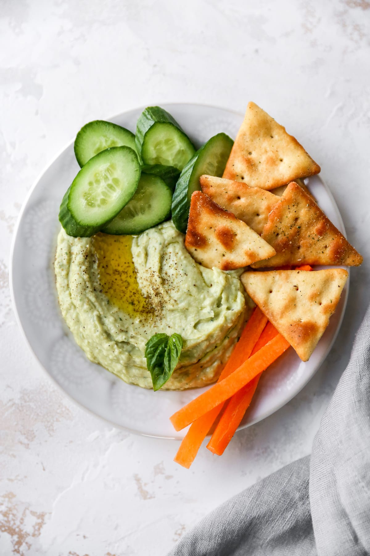 Bowl of lima bean hummus garnished with olive oil, pepper and basil and served with cucumbers, carrots and crackers.