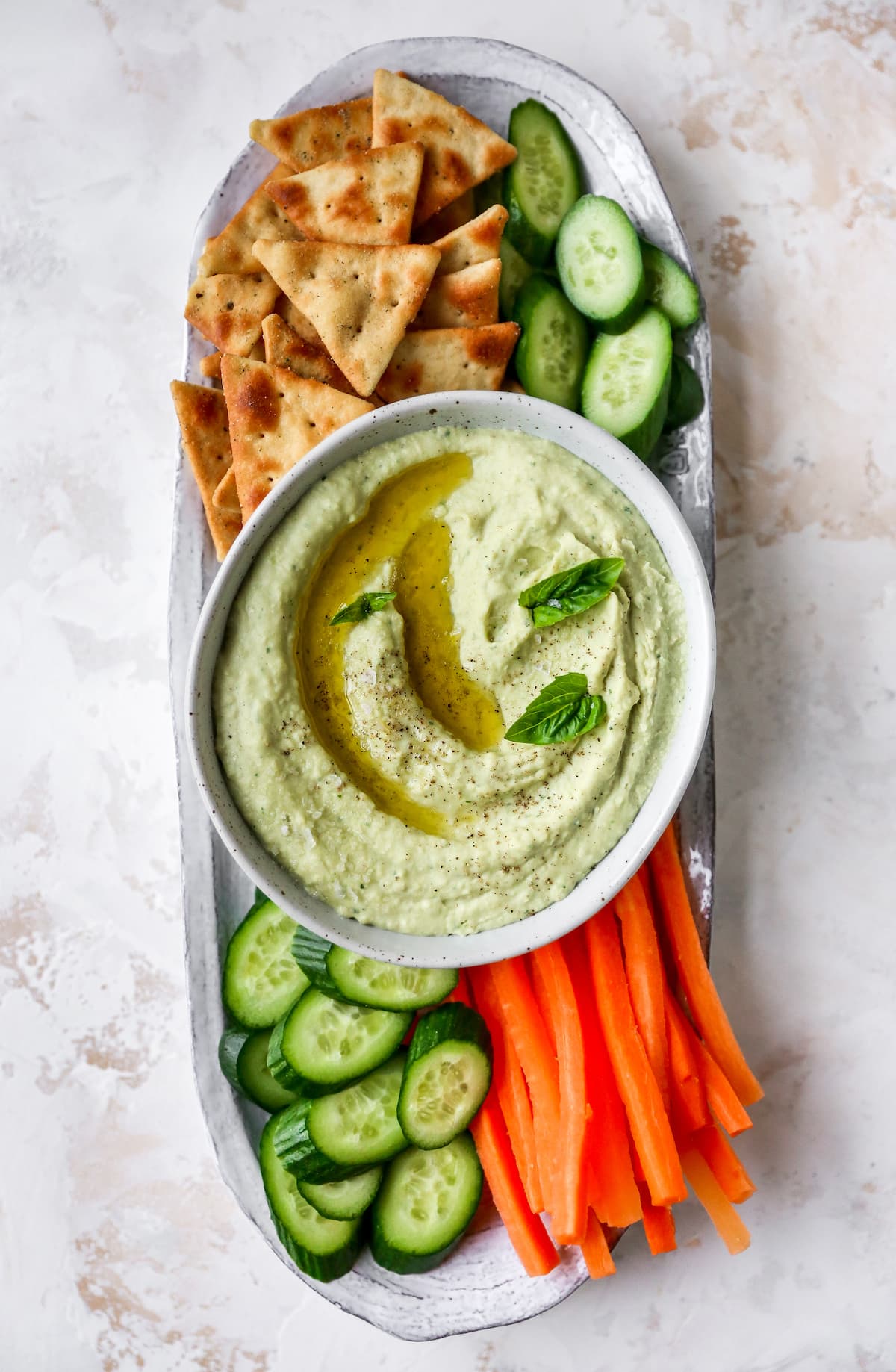 Lima bean hummus in a bowl garnished with basil, olive oil and pepper. Bowl is on a snack plate with cucumbers, carrots and crackers.