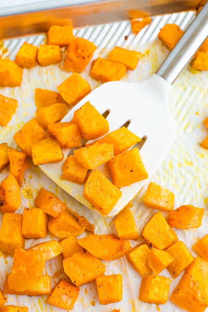 Spatula lifting up some roasted butternut squash cubes from a sheet pan.