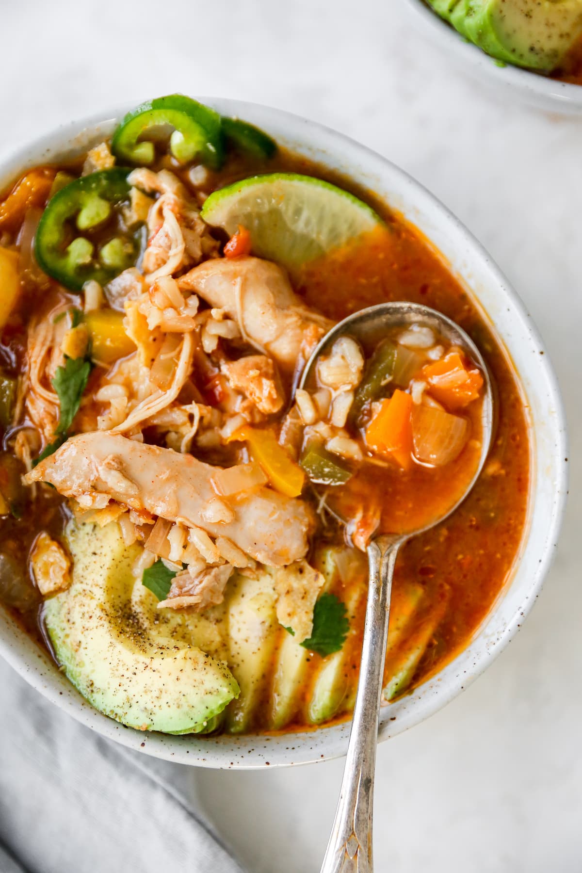 Bowl of chicken tortilla soup with avocado, line, jalapeno and chips as garnish. Spoon is in the bowl of soup.