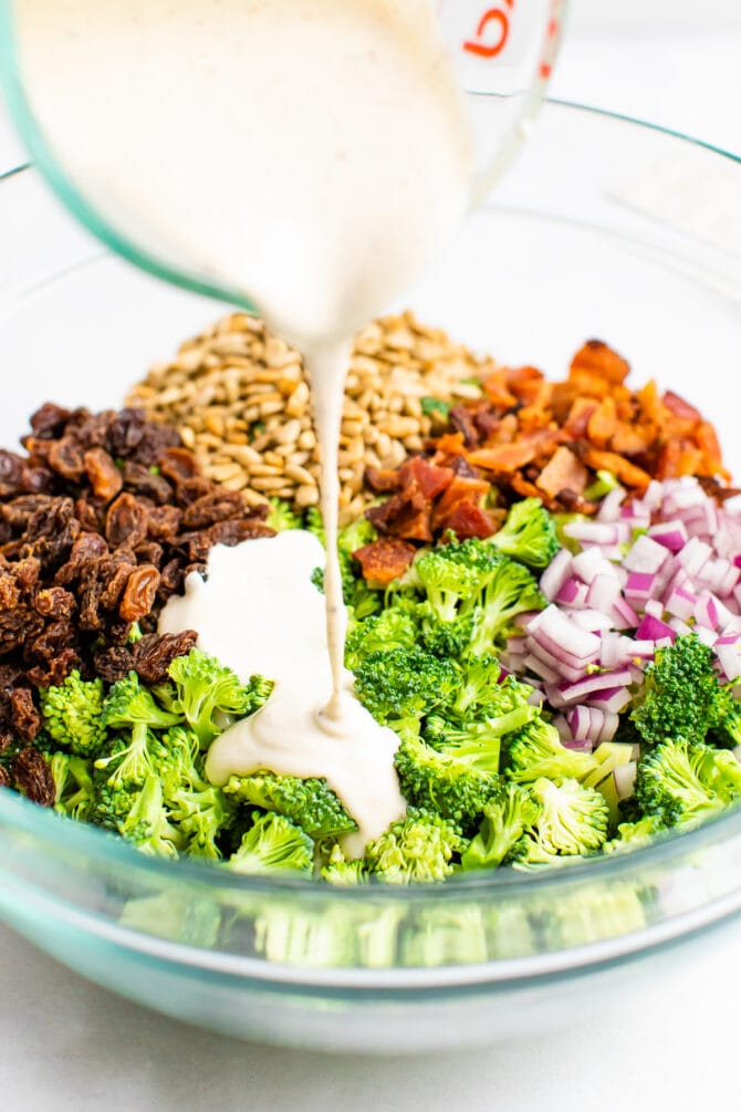 Creamy dressing being poured over a salad bowl of broccoli, onion, bacon, sunflower seeds and raisins.