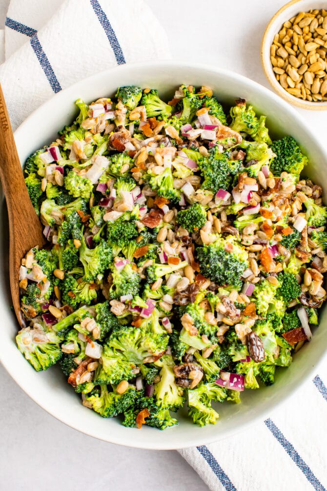 Bowl of broccoli salad with a wood spoon. A bowl of sunflower seeds is beside the bowl.