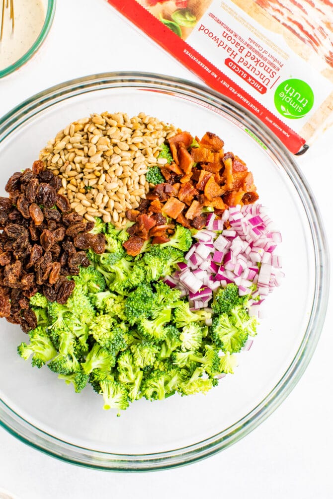 Glass bowl with broccoli, raisins, sunflower seeds, bacon and red onion. A package of bacon is next to the bowl.