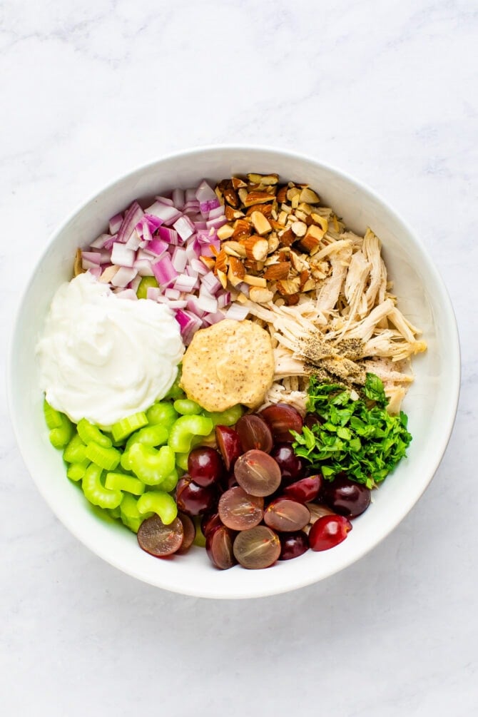 Mixing bowl with ingredients for Greek yogurt chicken salad before being mixed. Ingredients in the bowl are grapes, celery, parsley, greek yogurt, red onion, almonds and chicken.