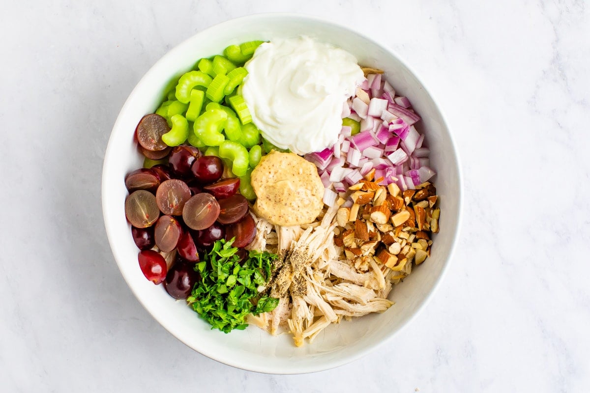 Mixing bowl with ingredients for Greek yogurt chicken salad before being mixed. Ingredients in the bowl are grapes, celery, parsley, greek yogurt, red onion, almonds and chicken.