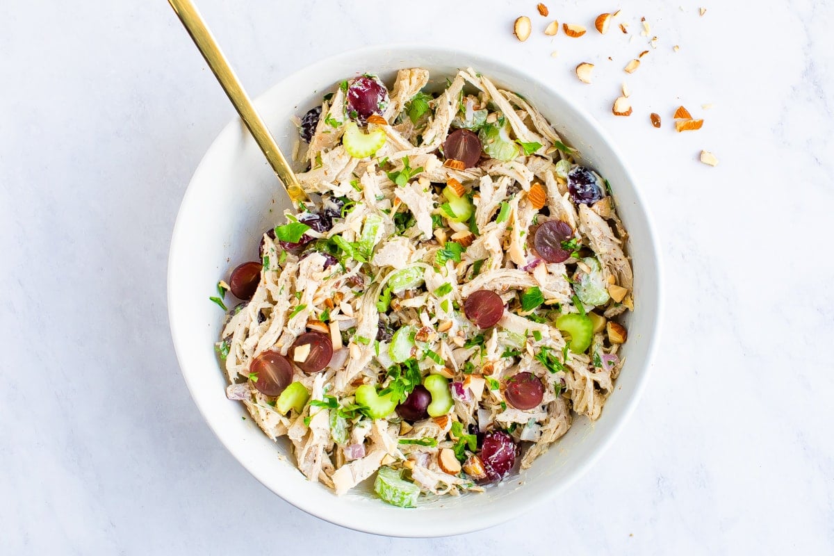 Mixing bowl with a spoon stirring chicken salad studded with celery and grapes.