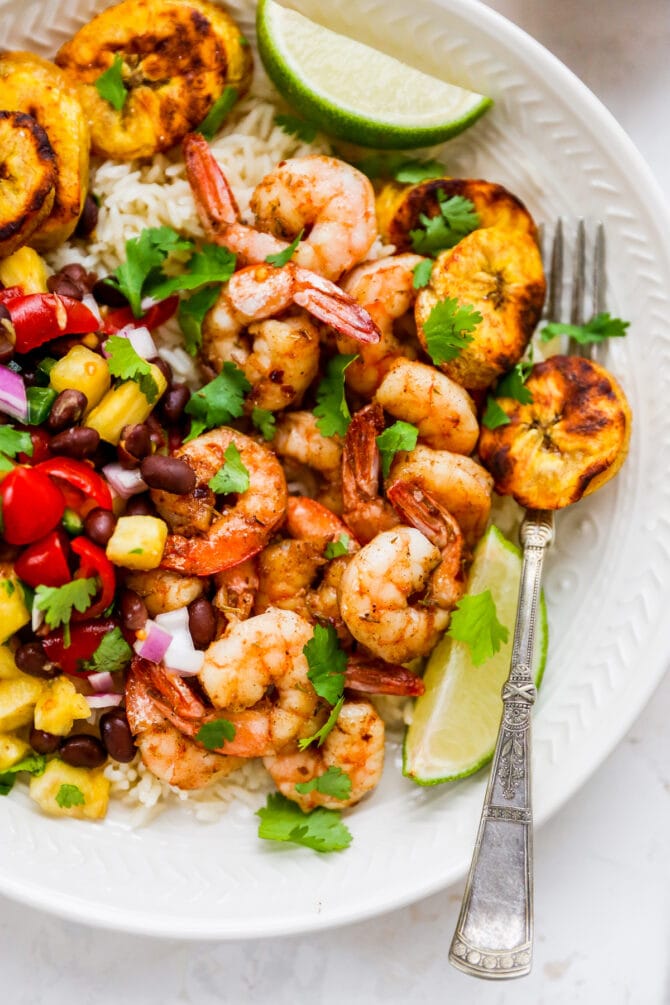 Plate with rice, pineapple salsa, jerk shrimp and plantains with lime and cilantro for garnish. Fork is on the plate.