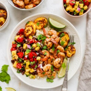 Plate with rice, pineapple salsa, jerk shrimp and plantains with cilantro and lime for garnish. A bowl of shrimp and pineapple salsa are beside the plate.