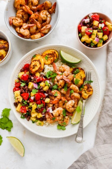 Plate with rice, pineapple salsa, jerk shrimp and plantains with cilantro and lime for garnish. A bowl of shrimp and pineapple salsa are beside the plate.