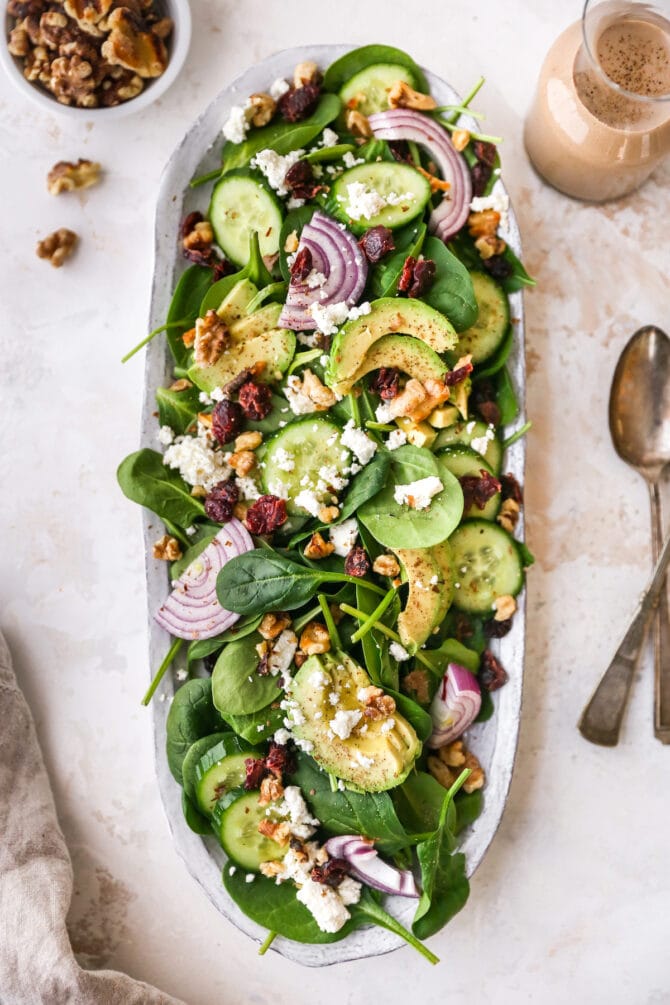 Spinach salad on a serving platter with cheese, nuts, onions and cranberries. Utensils, a bowl of walnuts and a jar of dressing are beside the tray.