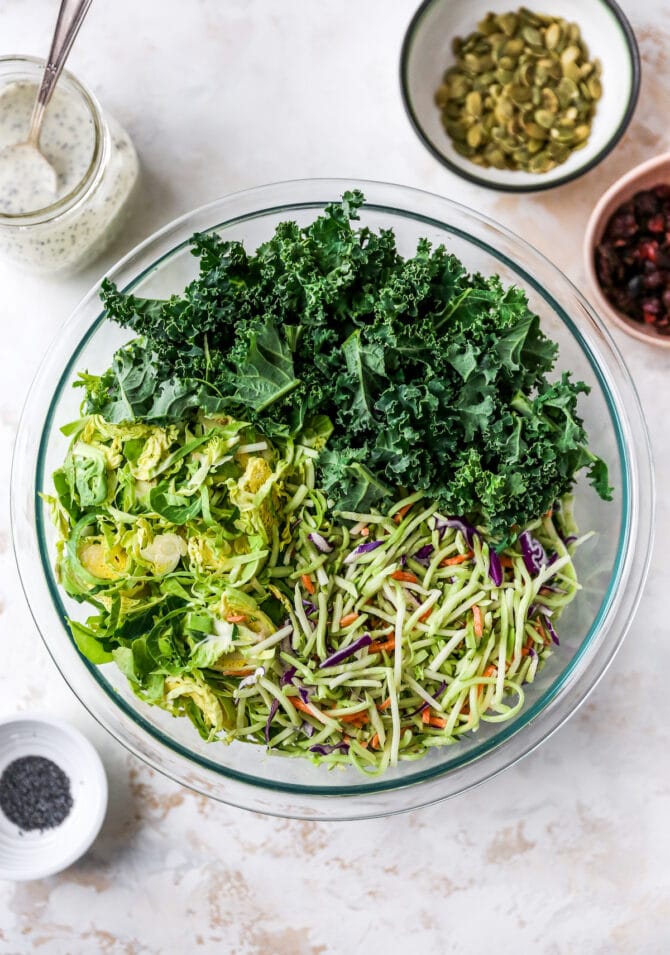Glass mixing bowl with broccoli slaw, brussels and kale. Dressing, pepitas, poppyseeds and cranberries are beside the bowl.