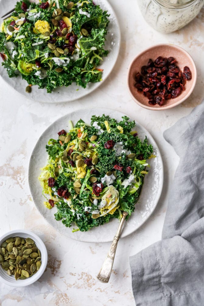 Plates of sweet kale salad with bowls of pepitas and cranberries on the side.