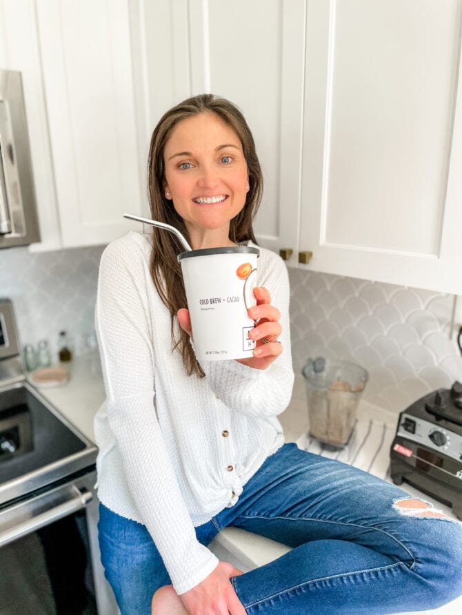 Woman sitting on a kitchen counter, holding a Daily Harvest smoothie and smiling.