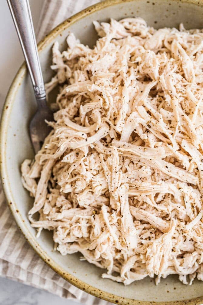 A close up of shredded chicken in a shallow bowl resting on top of a striped dish towel. A fork rests inside of the bowl.