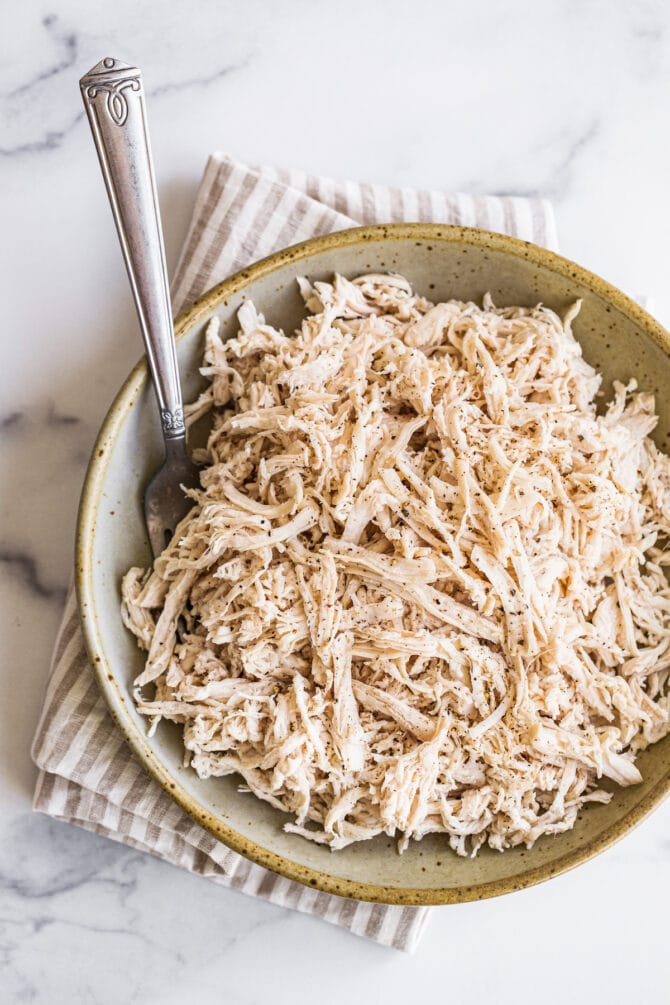 Shredded chicken in a shallow bowl resting on top of a striped dish towel. A fork rests inside of the bowl.