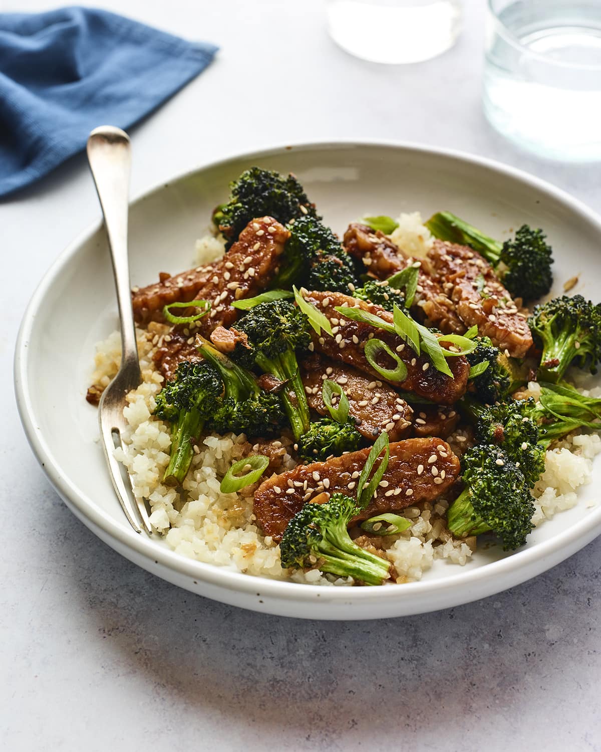 Bowl with rice topped with teriyaki tempeh and broccoli and sesame seeds. A fork is in the bowl.