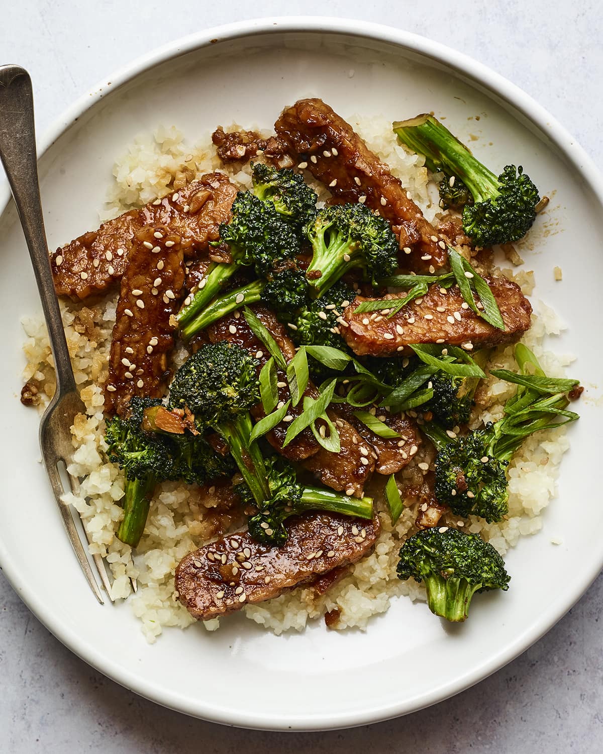 Bowl with rice topped with teriyaki tempeh and broccoli and sesame seeds. A fork is in the bowl.