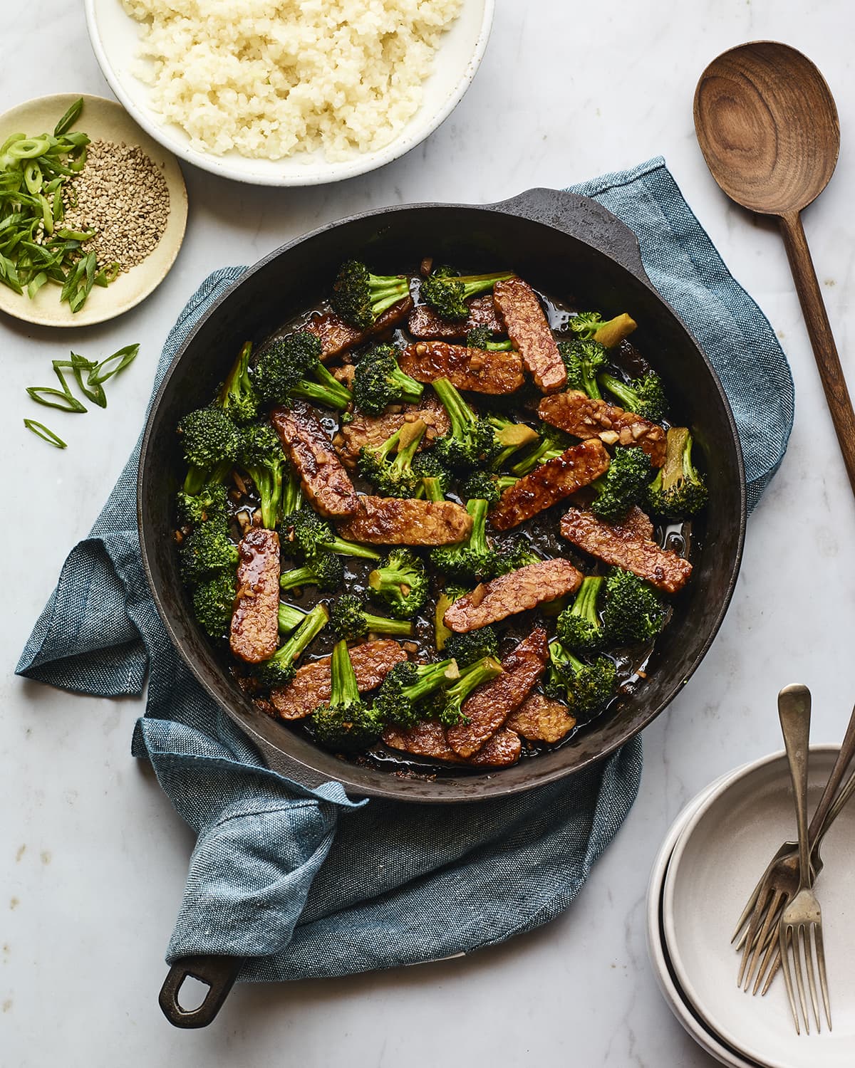 Teriyaki tempeh and broccoli in a cast iron skillet. A cloth towel is under the skillet. A bowl of rice, a wood spoon and bowls and forks are beside the skillet.