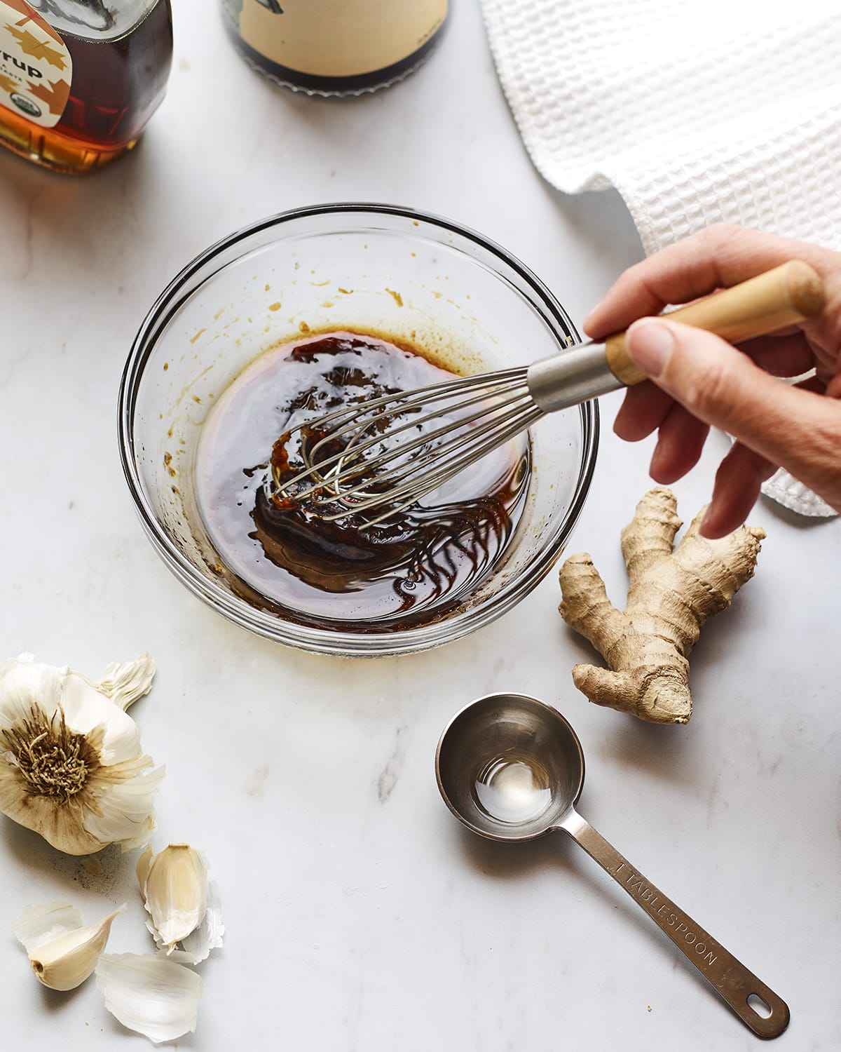 Hank whisking together teriyaki sauce in a glass bowl. Garlic, ginger, a measuring spoon and a bottle of maple syrup are on the counter.