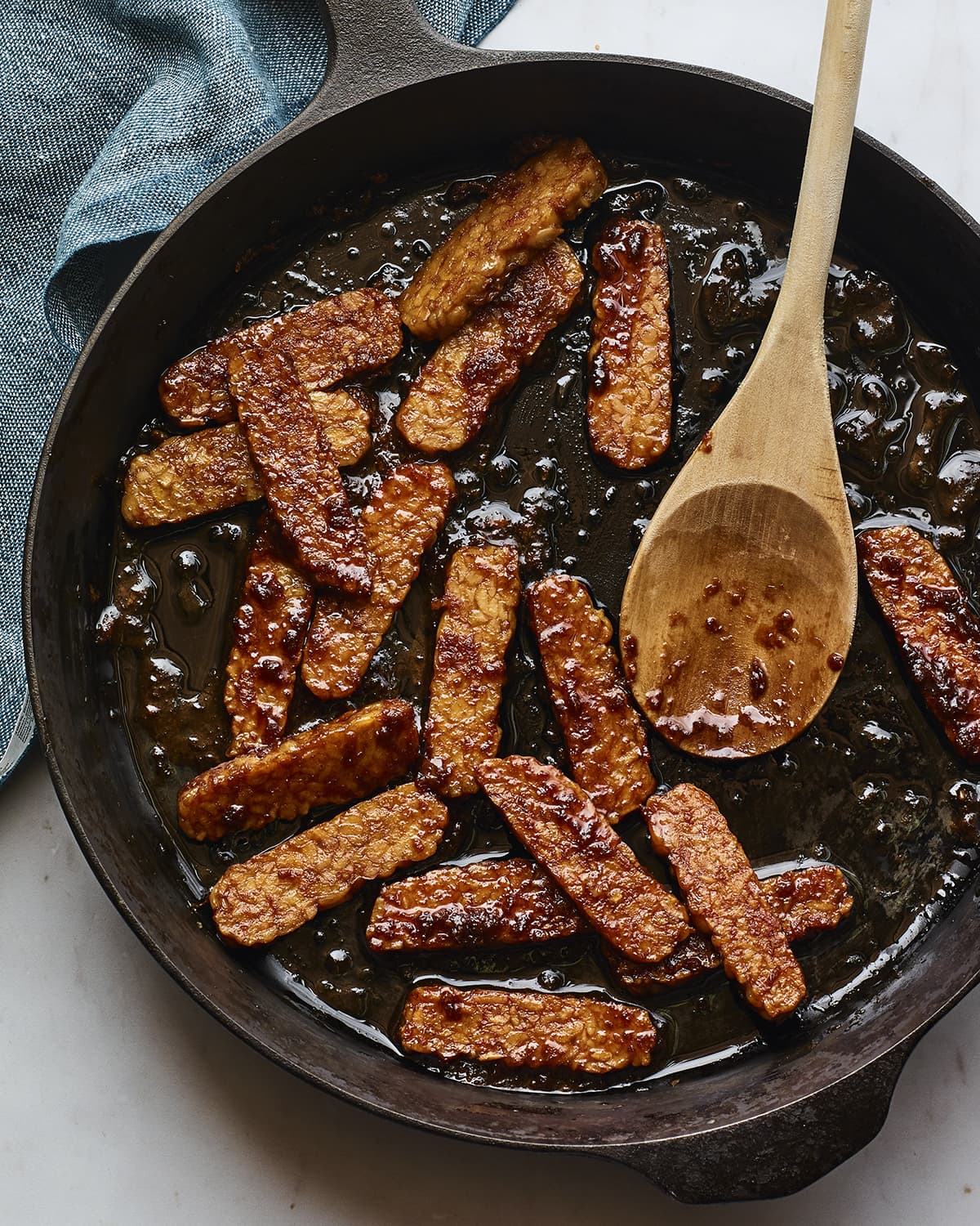 Pieces of teriyaki tempeh cooking in a cast iron skillet. A wooden spoon is in the skillet.