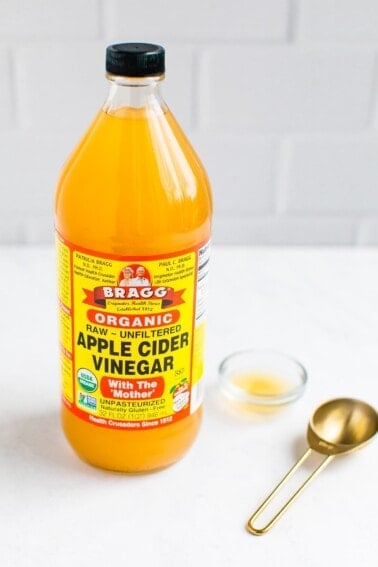 A bottle of Bragg Organic Apple Cider Vinegar with a gold tablespoon on the side.