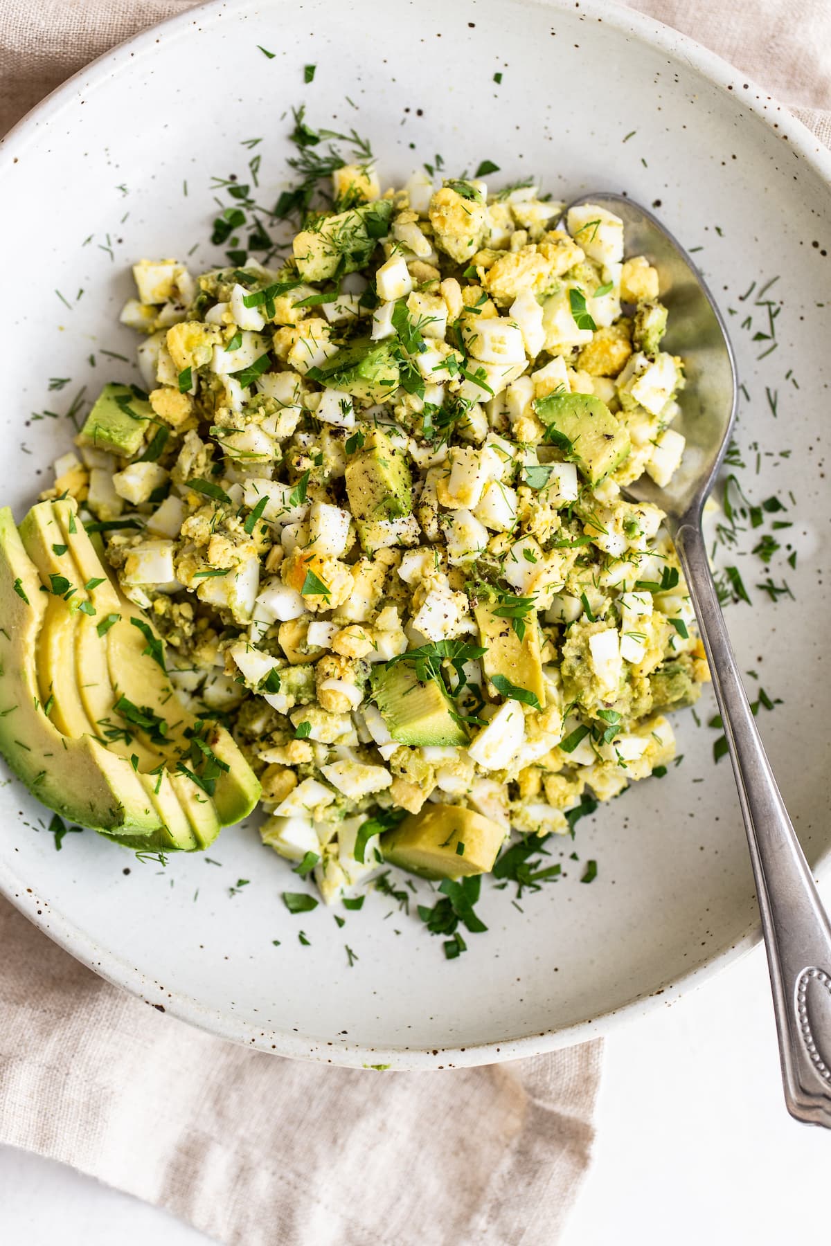 Bowl with avocado egg salad and spoon. Salad is garnished with avocado slices and herbs.