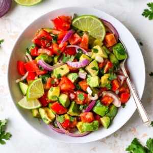 Serving bowl with avocado and tomato salad garnished with lime and cilantro. A spoon is in the bowl.