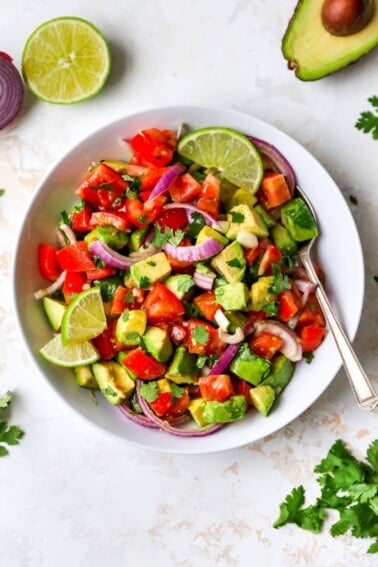 Serving bowl with avocado and tomato salad garnished with lime and cilantro. A spoon is in the bowl.