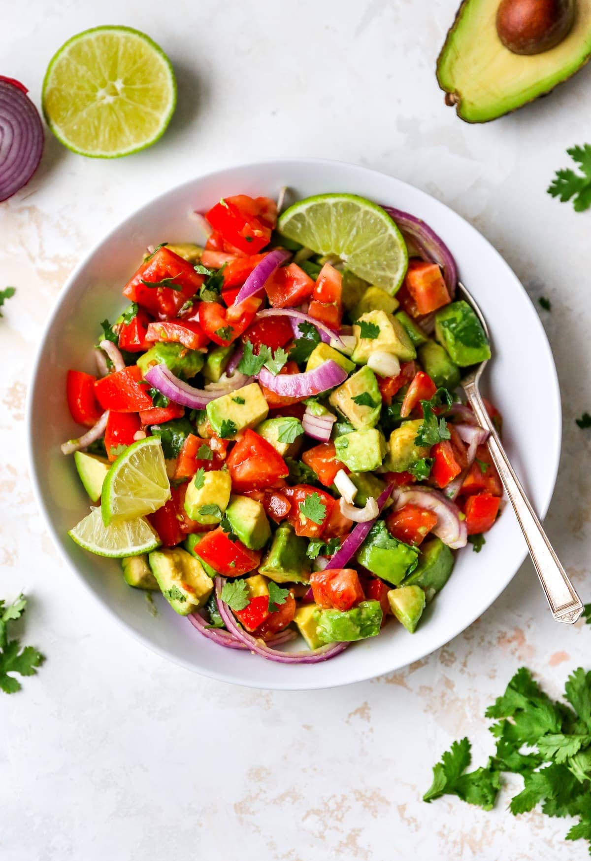 Serving bowl with avocado and tomato salad garnished with lime and cilantro. A spoon is in the bowl.