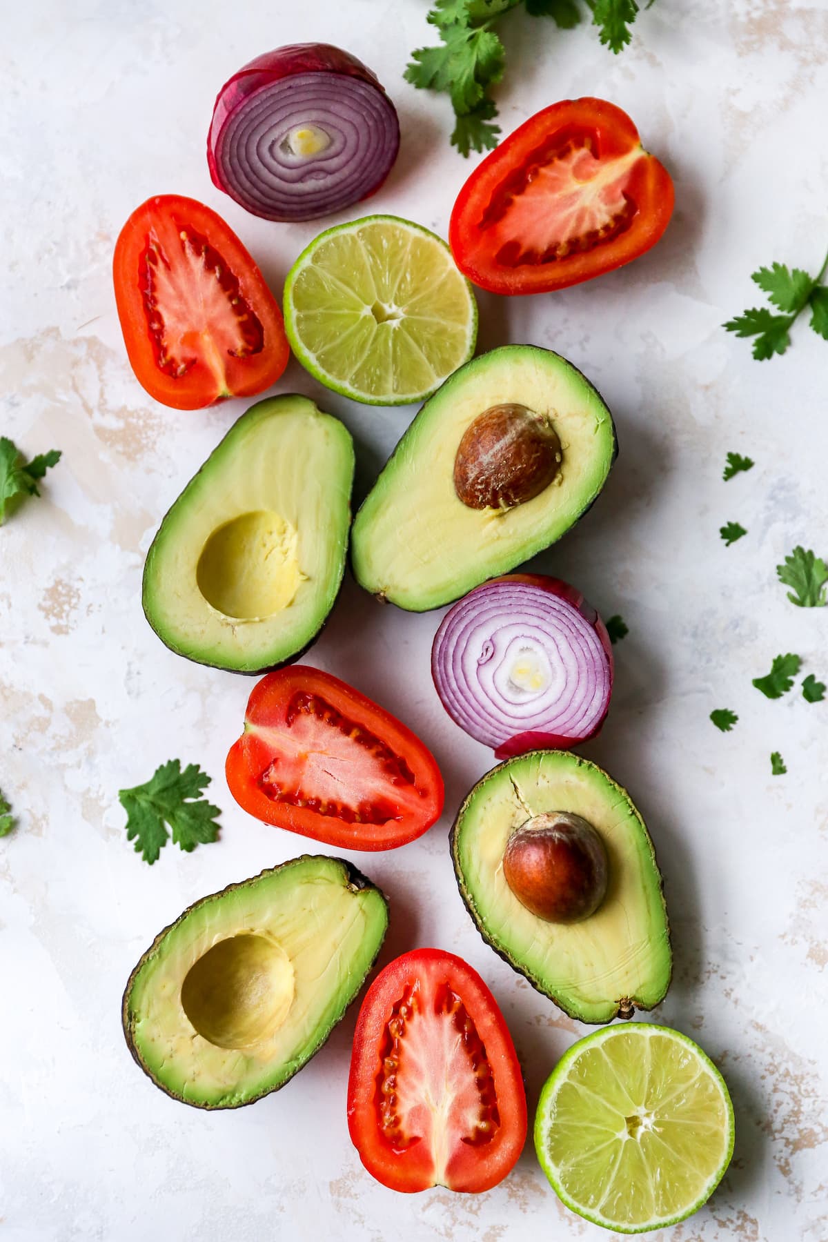 Halves of red onion, avocado and tomatoes on a table surrounded by fresh cilantro.