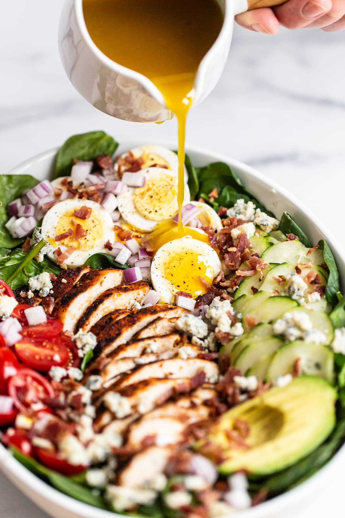 Pouring red wine vinaigrette dressing over a cobb salad made with spinach, egg, onion, bacon, blue cheese, cucumber, avocado, blackened chicken and tomatoes.