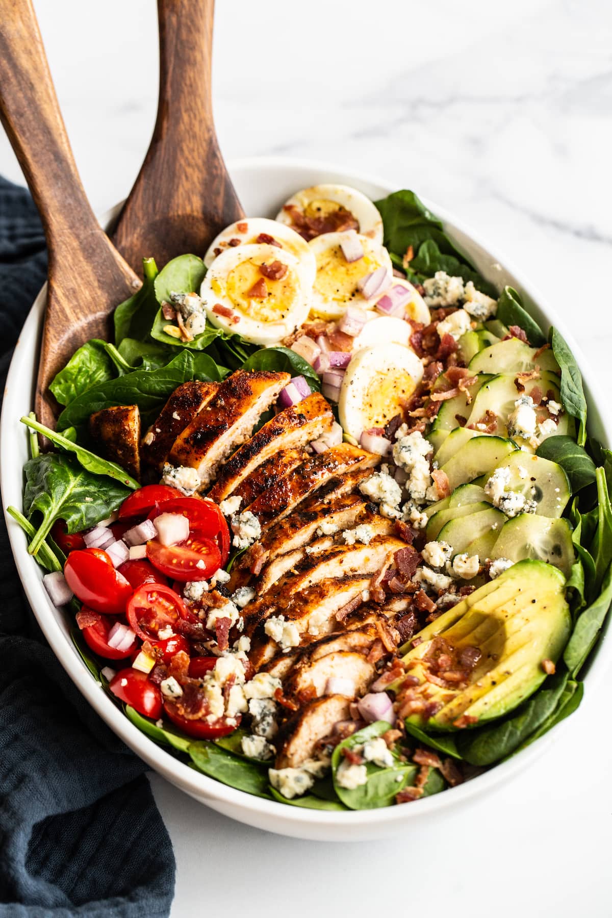 Cobb salad in a large serving bowl with hard boiled egg, blue cheese, cucumber, avocado, blackened chicken, tomato, spinach and bacon. Two wood salad serving spoons are in the bowl.